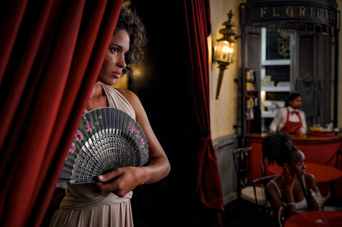 A woman holds an open fan beside curtains, with other people seated in the room behind her.