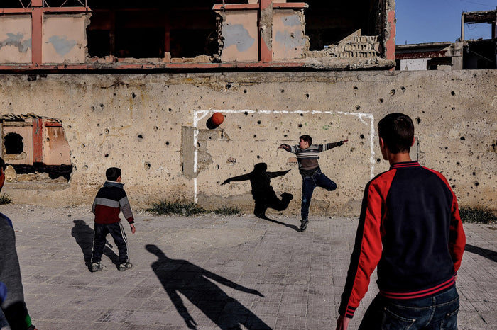 Children play football in front of a damaged wall, with one child leaping toward a ball.